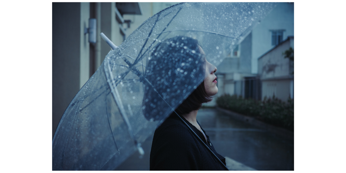 Woman in dark clothing holding clear umbrella with raindrops on it looks up.