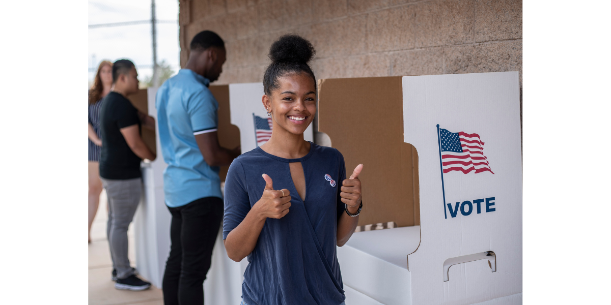 Woman standing in front of voting kiosk holding two thumbs up. Others in background fill out their ballots.